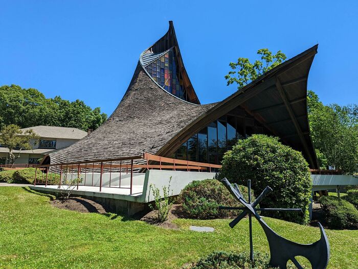 Unique bizarre building with curved wooden roof and large glass windows surrounded by greenery under a clear blue sky.