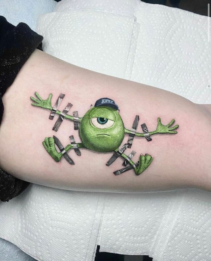 Crazy tattoo of a green cartoon character with one eye and limbs outstretched, inked on an arm.