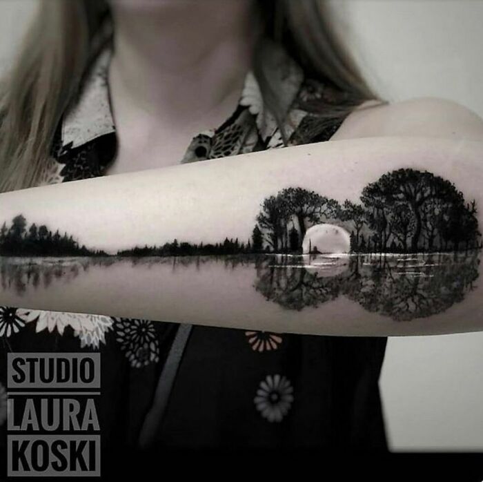 Arm with landscape tattoo featuring trees and a moon reflected in water.