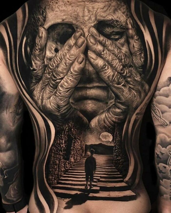 Intricate back tattoo depicting hands covering a face with a surreal landscape scene.