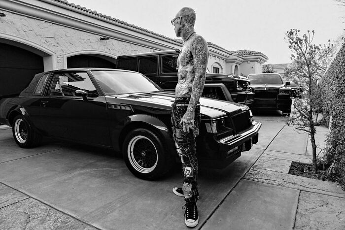 Tattooed celebrity posing with luxury cars, showcasing wealth in a driveway.