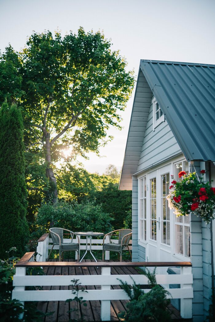 Cozy wooden porch with chairs and table by a vintage house surrounded by lush green trees on a sunny day.