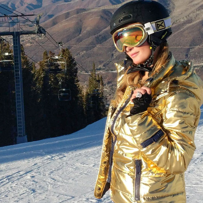 Celebrity flaunting wealth on a ski slope in a shiny gold jacket, wearing a helmet and reflective goggles.