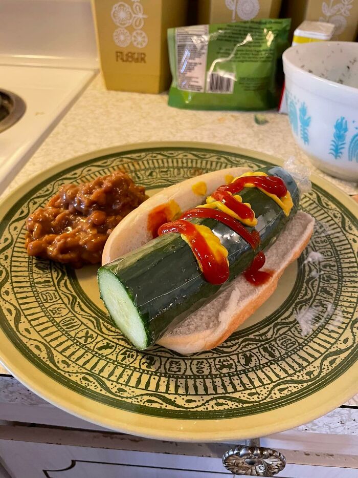 Unusual food example with whole cucumber in a hot dog bun topped with ketchup and mustard, served with baked beans.