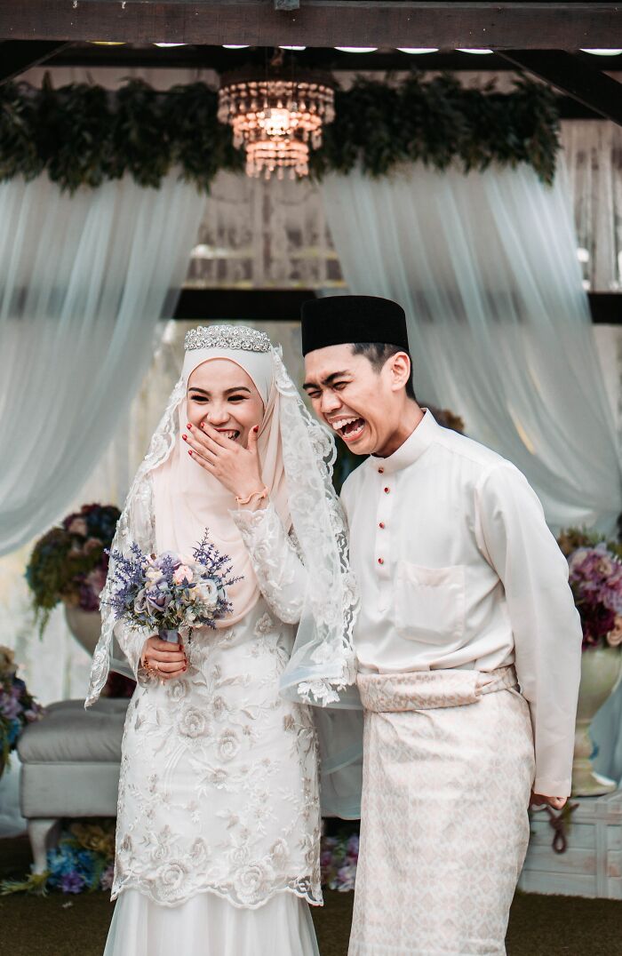 Bride and groom in traditional attire laughing joyfully during a unique wedding experience with floral decor and draped curtains.