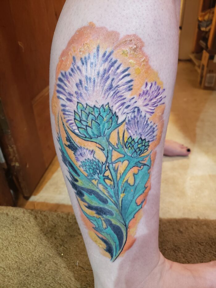 Colorful floral tattoo on a leg featuring detailed thistle flowers.