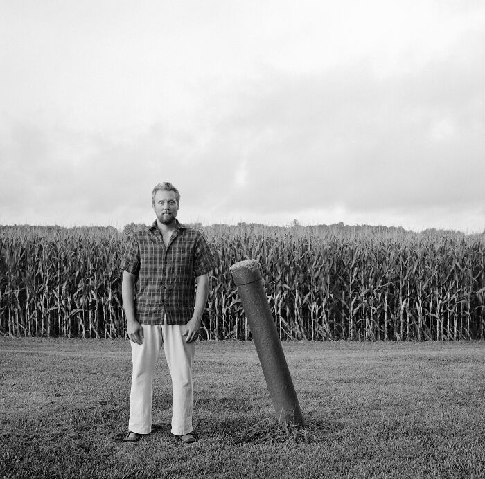 Black and white powerful portrait of a man standing near a tilted post in front of a cornfield under cloudy sky.