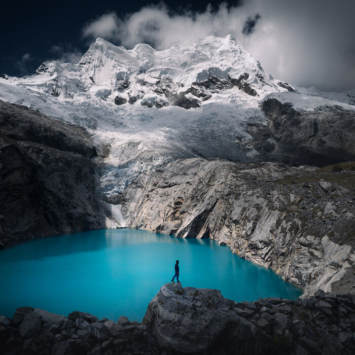 A person stands on rocks by a turquoise lake in the Peruvian Andes with snowy mountain peaks and clouds in the background.
