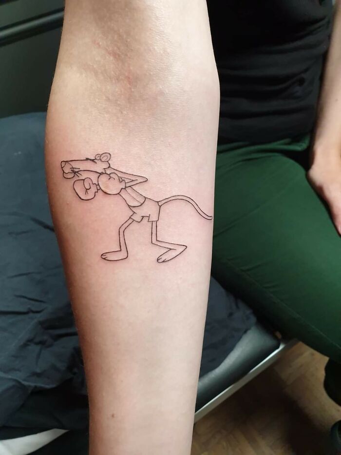 Tattoo of a cartoon character on a forearm, featuring a playful pose, displayed by a person in green pants.