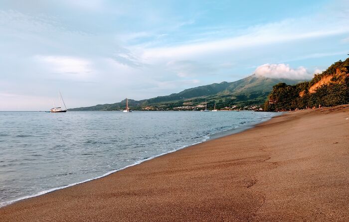 A French Caribbean Island. Cliff On The Right, Volcano In The Back, The Caribbean Sea On The Left And Sweet Sands Under The Feets