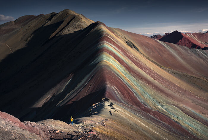 Colorful mountain ridges in the Peruvian Andes with a lone traveler standing at the base during clear weather.