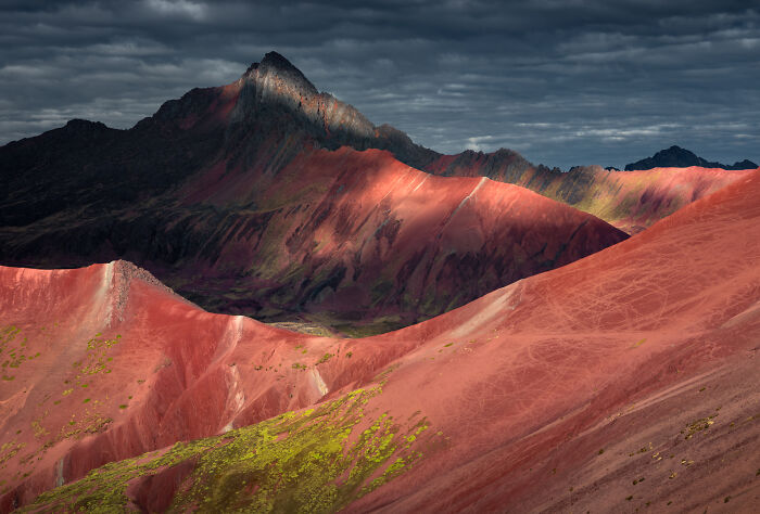 Vibrant red and green slopes of the Peruvian Andes under dramatic clouds with sunlight highlighting the mountain peaks.