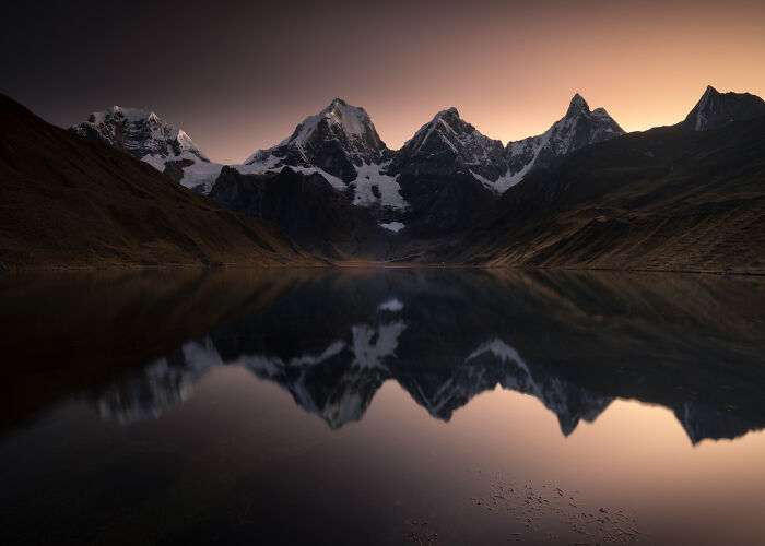 Snow-capped peaks of the Peruvian Andes reflected in a calm mountain lake during sunset with warm orange sky.