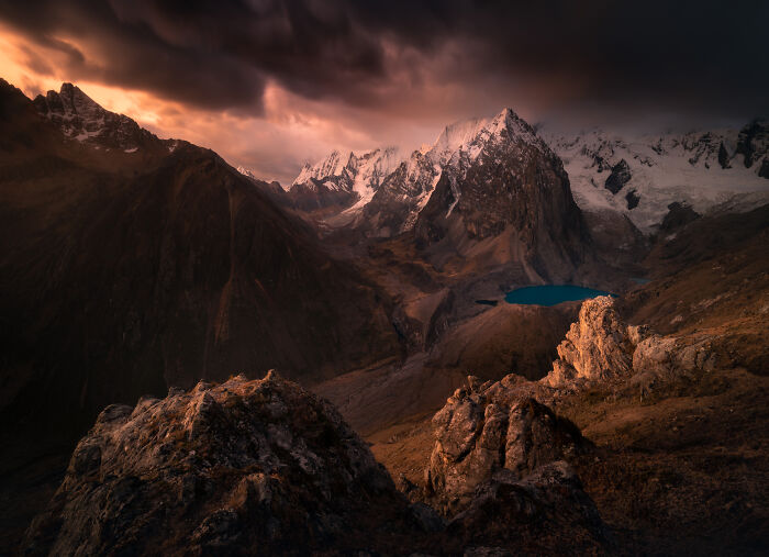 Dramatic sunset over the Peruvian Andes mountains with rugged peaks, rocky foreground, and a small turquoise lake below.