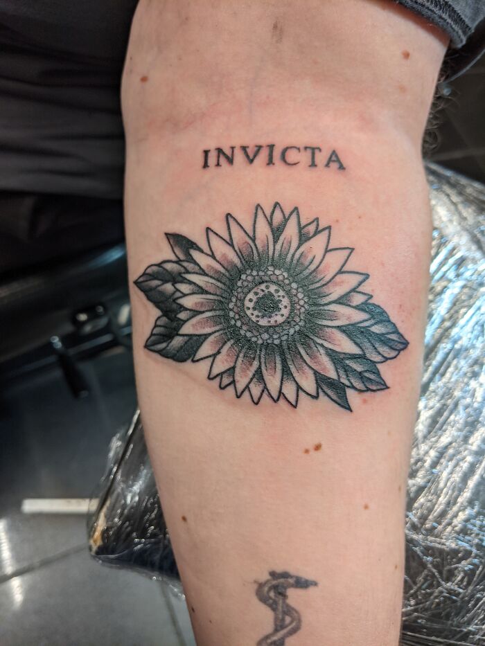 Sunflower tattoo with "INVICTA" text on forearm, featuring detailed leaves and petals.