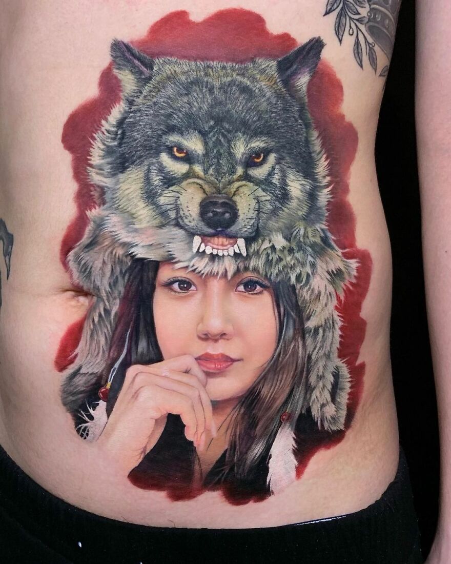 Hyper-realistic tattoo of a woman's face with a wolf headdress, intricately detailed on skin.