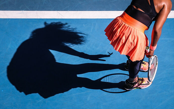 Tennis player in an orange skirt ready to serve, casting a dramatic shadow on the blue court, showcasing sports moments.