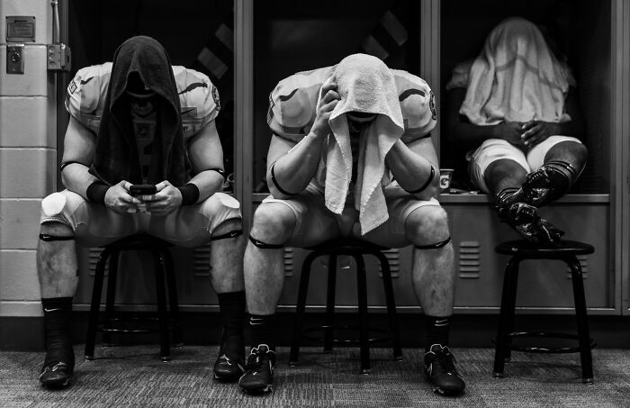 Three football players with towels over their heads sitting in a locker room capturing intense moments in sports photography.