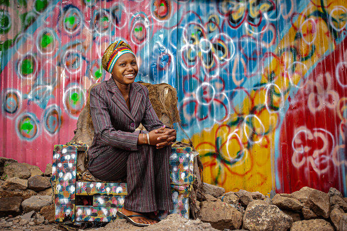 Smiling woman in colorful headwrap and striped suit sitting on a painted chair with vibrant graffiti in powerful portrait.