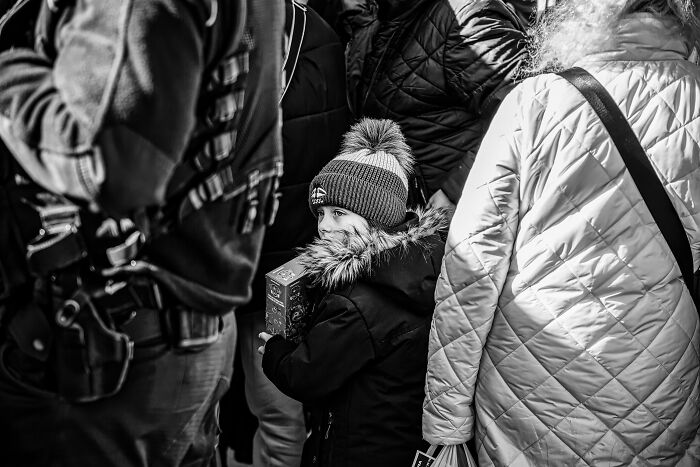Black and white portrait of a young child in winter clothes holding a box in a crowded street scene by photographers