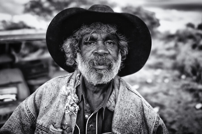 Black and white powerful portrait of an elderly man with a beard wearing a wide-brimmed hat and denim jacket outdoors