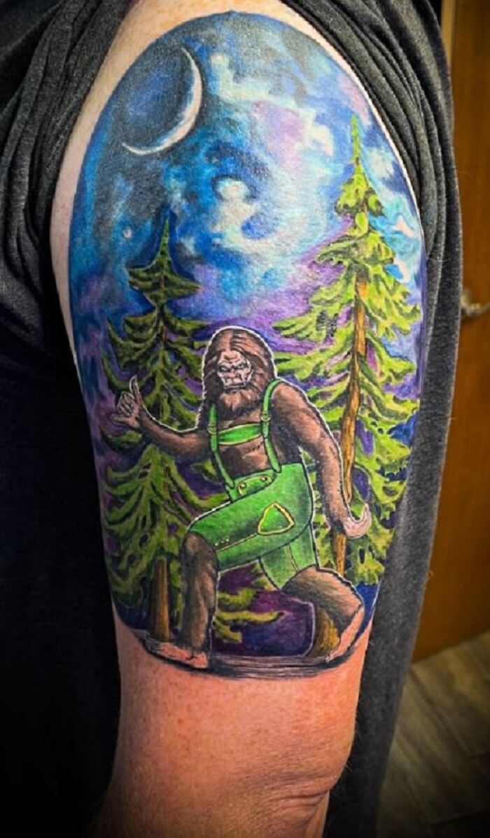 Tattoo featuring Bigfoot in green overalls walking through a forest under a crescent moon.