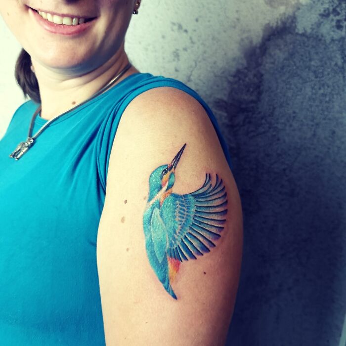 Person showcasing a colorful bird tattoo on their upper arm, wearing a blue shirt and smiling.