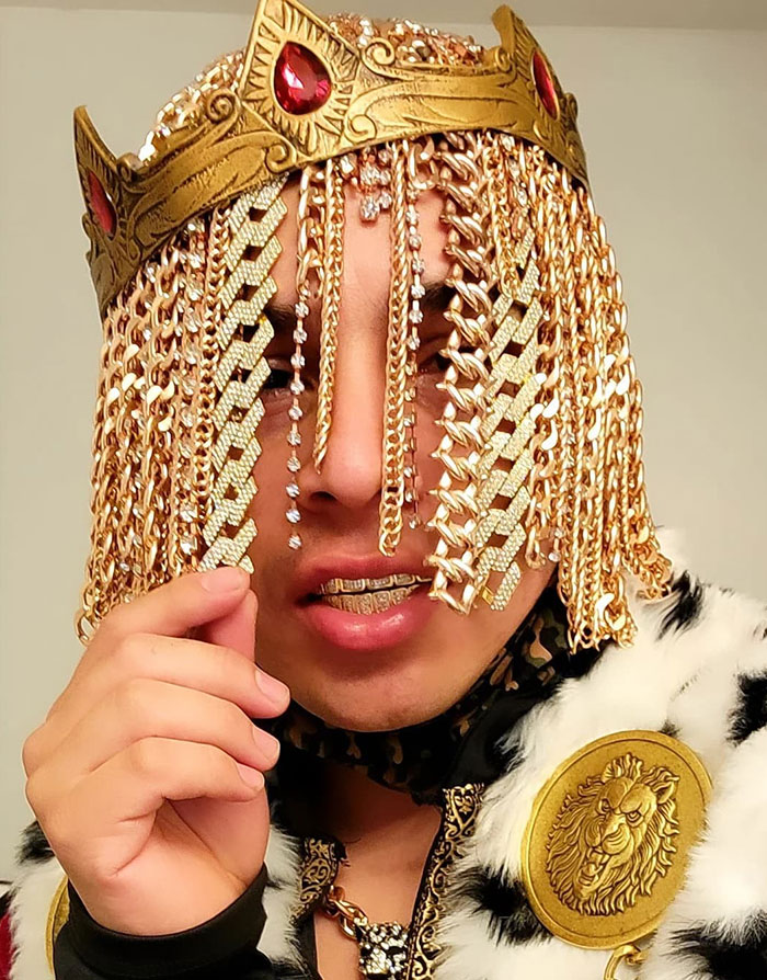 Celebrity flaunting wealth wearing a gold crown with jewels, gold chains, and a fur-trimmed robe.
