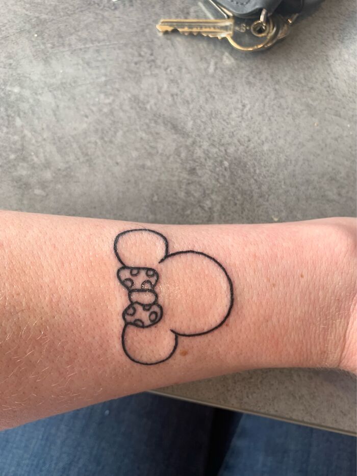 Tattoo of a bear face outline with a polka dot bow on an arm next to keys on a table.