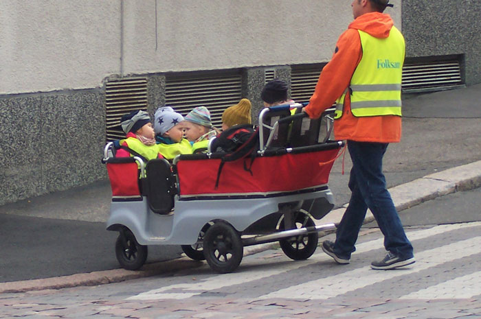 Adult in safety vest pushing stroller with multiple toddlers on city sidewalk, illustrating entertaining it doesn't work like that stories.