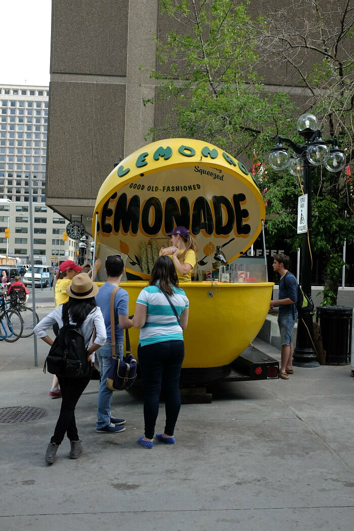 People buying lemonade at a vibrant yellow lemonade stand on a city sidewalk with trees and buildings nearby.