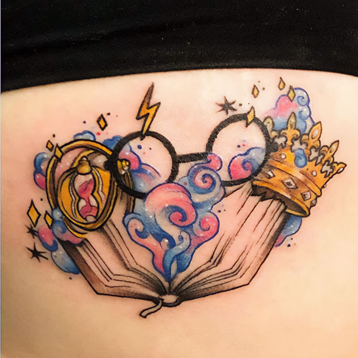 Tattoo of Harry Potter elements: glasses, a crown, a book, and a time-turner with colorful smoke and magic symbols.