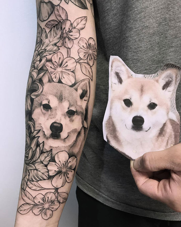 Pet tattoo of a dog's face surrounded by flowers on an arm, matching a photo held beside it.