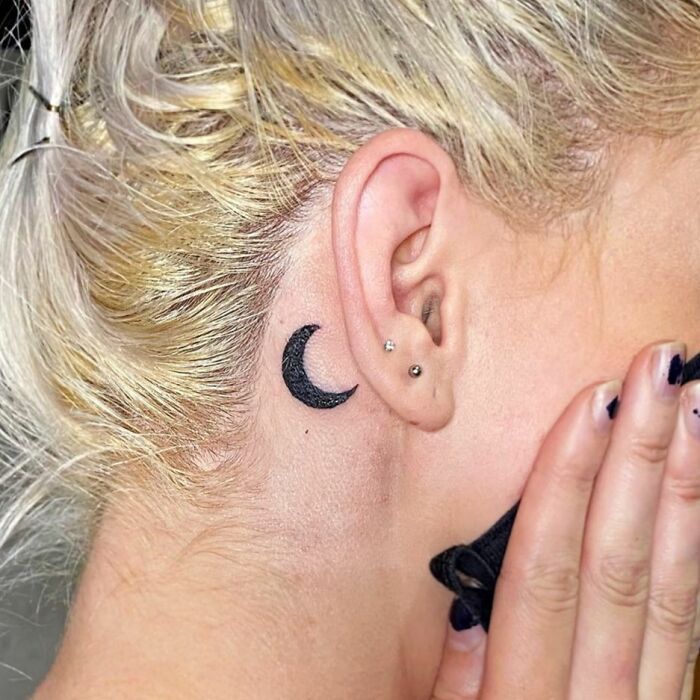 Ear tattoo featuring a black crescent moon behind the ear with small piercings, exemplifying subtle tattoo ideas.