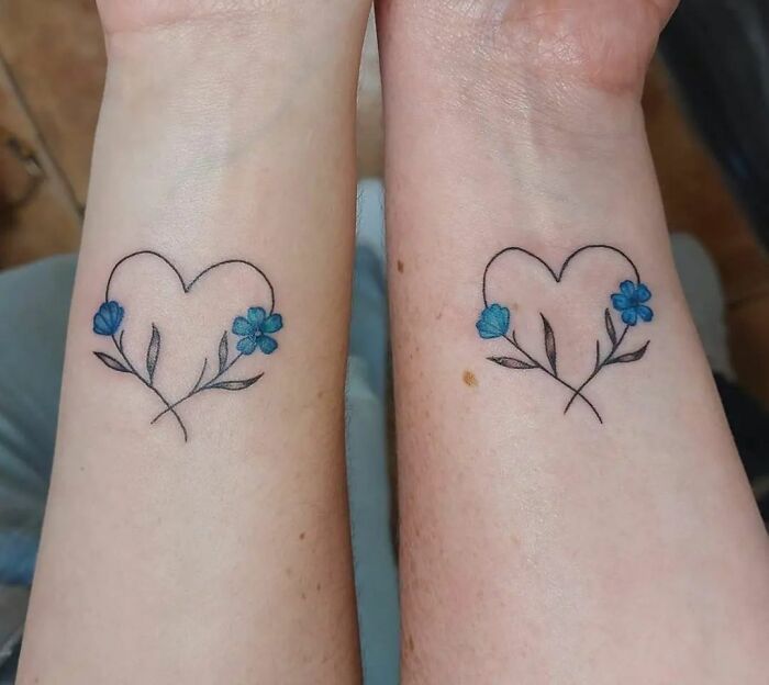 Matching heart tattoos with blue flowers on two wrists, symbolizing best friend tattoos for friendship.