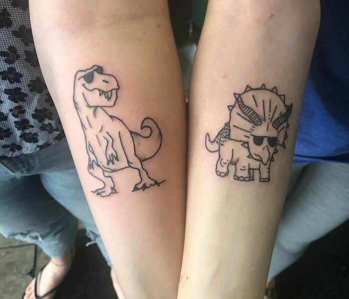 Two forearms with matching best friend tattoos of cartoon dinosaurs wearing sunglasses.