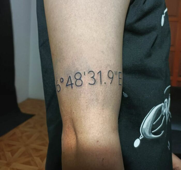 Armband tattoo featuring geographic coordinates in black ink on an arm.