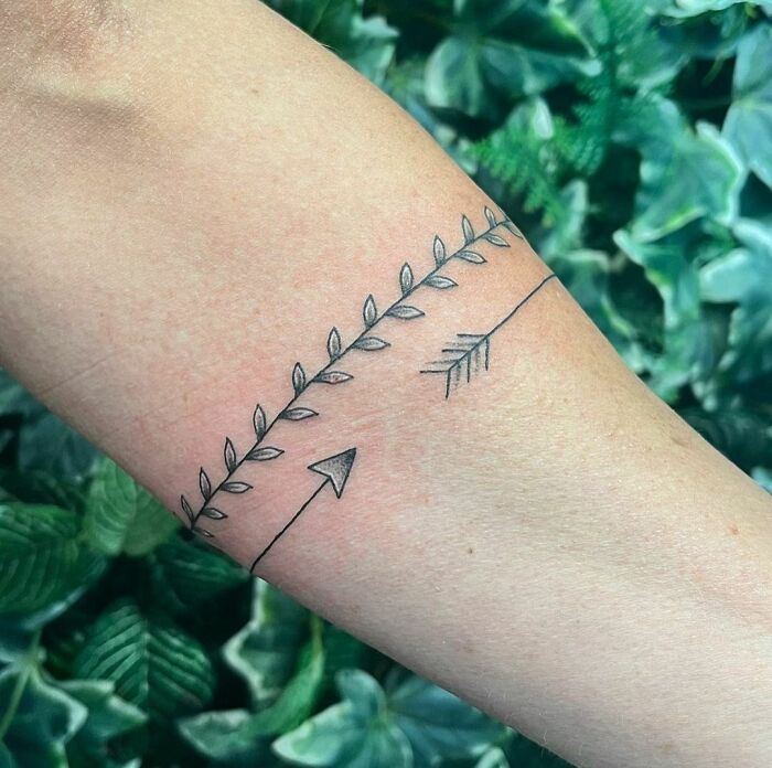 Minimalist armband tattoo featuring an arrow and leaf design against a green leafy background.