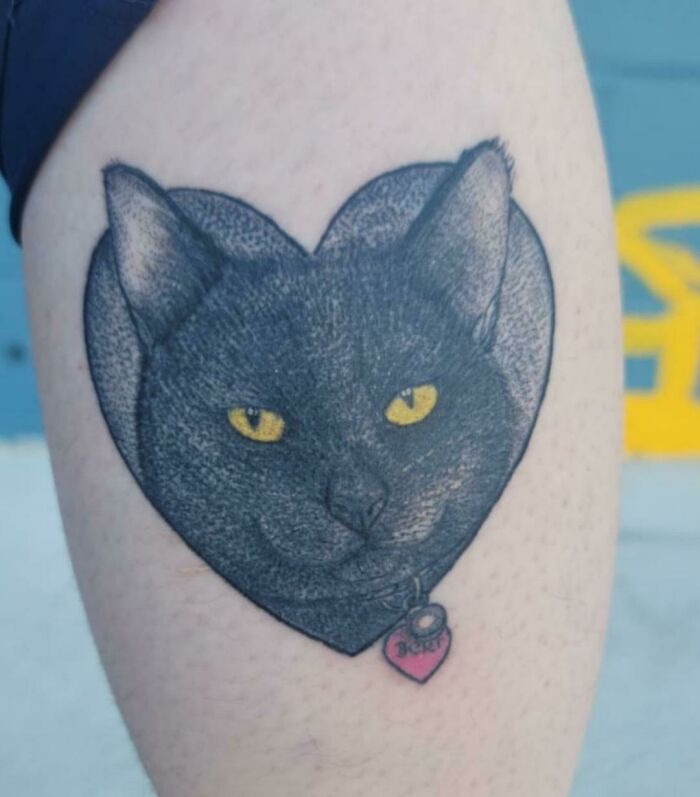 Heart-shaped black cat lower leg tattoo with yellow eyes and pink collar.