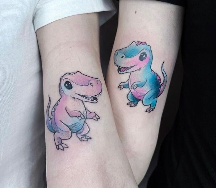 Cute dinosaur best friend tattoos on forearms, one pink and the other blue, symbolizing friendship.