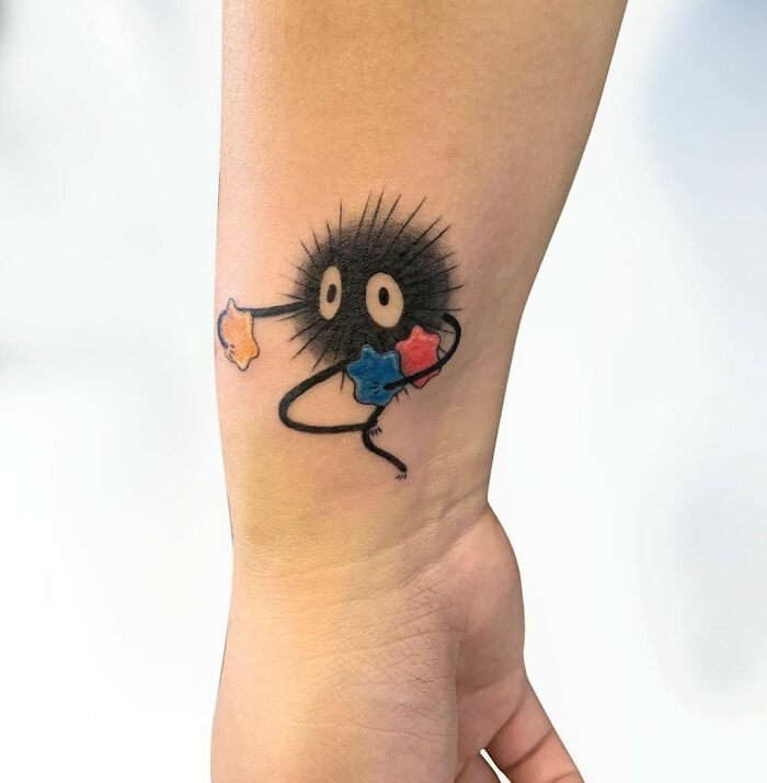 Cartoon tattoo of a soot sprite holding colorful stars on an arm.