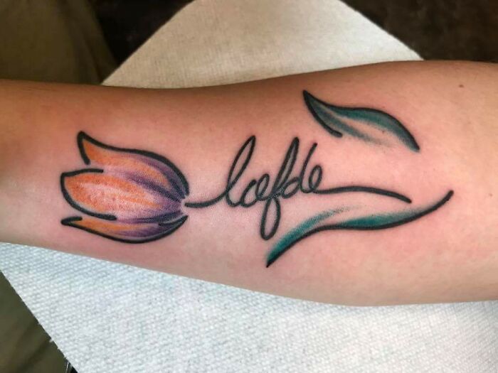 Floral tattoo with script on arm, featuring purple and orange flower with green leaves; meaningful word tattoos.
