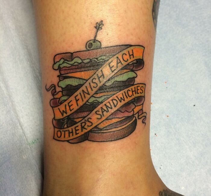 Tattoo design of a sandwich with text ribbons that read, "We finish each other's sandwiches," on skin.
