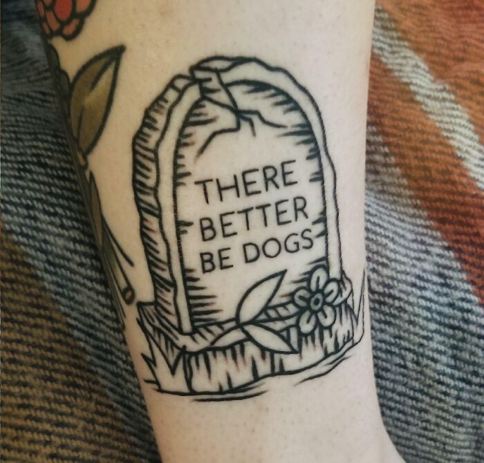 Creative word tattoo of a tombstone with "There better be dogs" on a person's arm, featuring leaves and flowers.