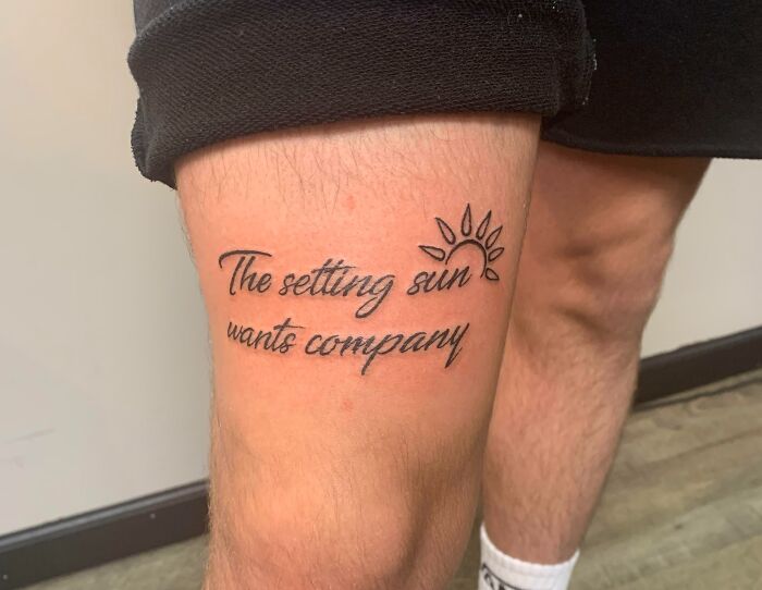 Word tattoo on a person's leg reading "The setting sun wants company" with a sun illustration.