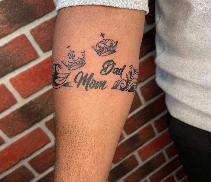 Armband tattoo with crowns and "Mom" and "Dad" lettering on forearm, set against a brick wall background.