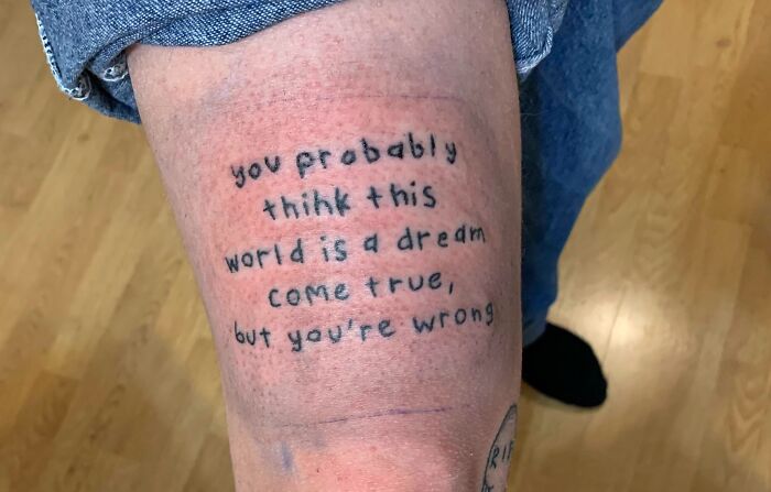 Arm tattoo reading "you probably think this world is a dream come true, but you're wrong" in simple black lettering.
