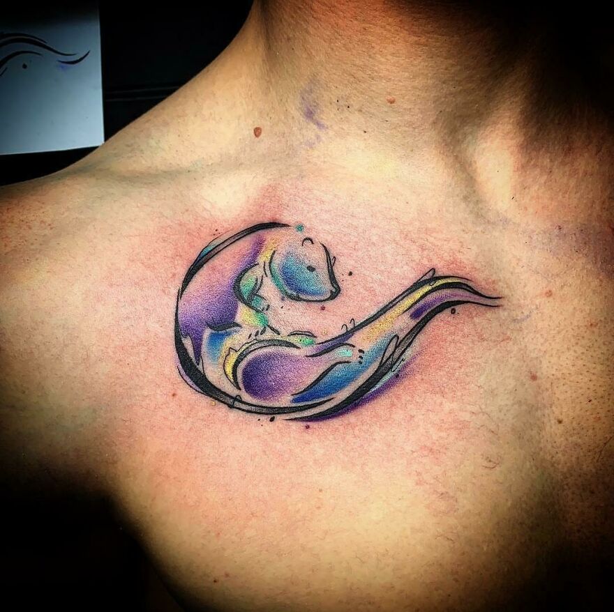 Elegant watercolor tattoo of an otter with vibrant purple and blue hues on a person's upper chest.