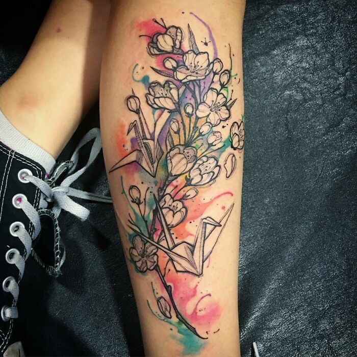 Watercolor tattoo with origami cranes and blossoms, featuring vibrant splashes on an arm.