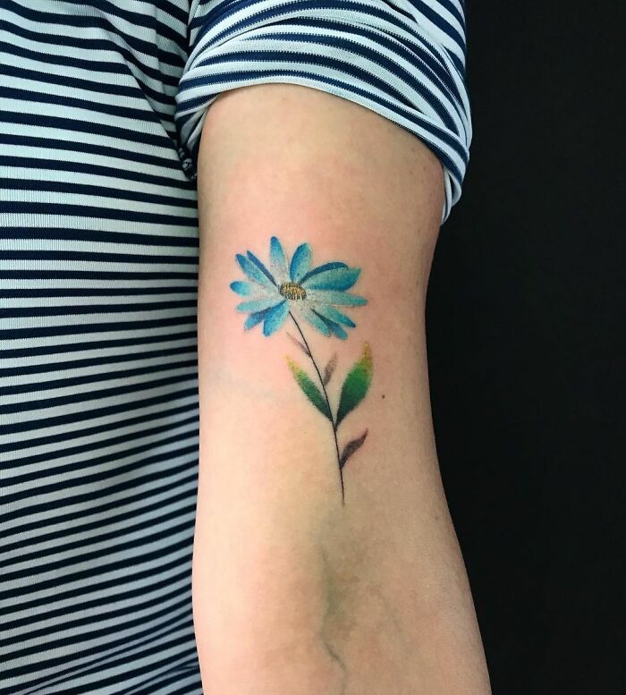 A watercolor tattoo of a blue flower on an arm, showcasing vibrant ink inspiration.